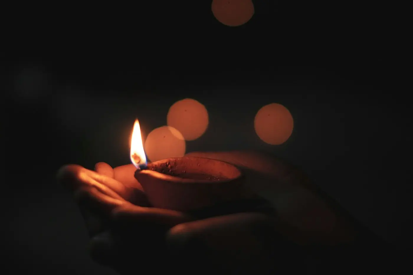 Two hands holding a lit clay diya in the dark